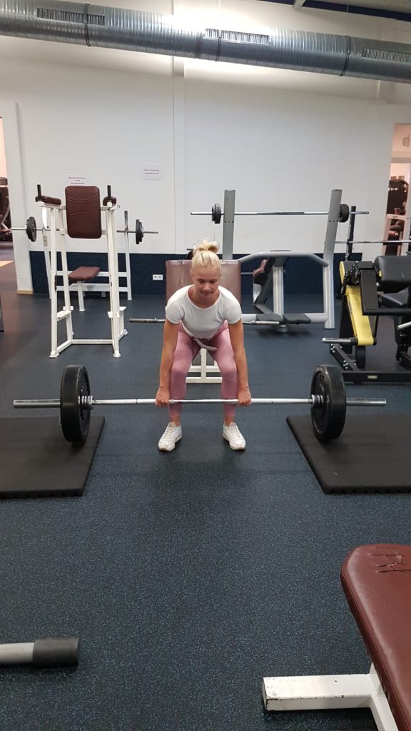 Person hebt Langhantel im Fitnessstudio, trägt weißes Oberteil und rosafarbene Leggings.