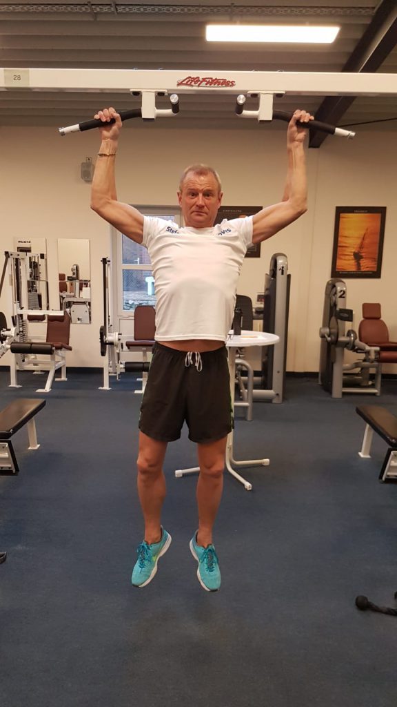 Person macht Klimmzüge an einer Stange im Fitnessstudio, umgeben von Trainingsgeräten.