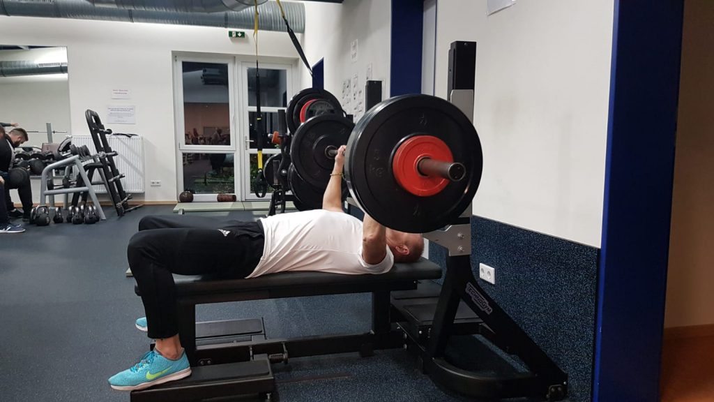 Person hebt Langhantel auf Flachbank im Fitnessstudio, umgeben von Geräten und fokussierter Atmosphäre.