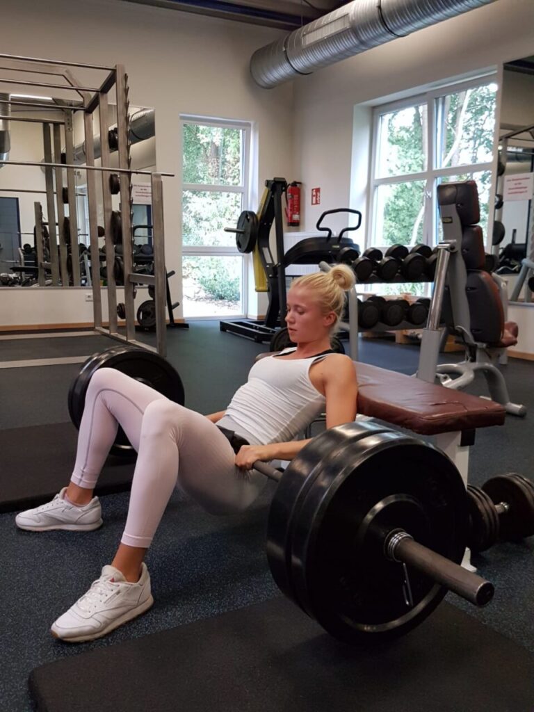 Frau im Fitnessstudio macht Hip Thrusts mit Langhantel auf gepolsterter Bank, trägt weiße Sportkleidung.