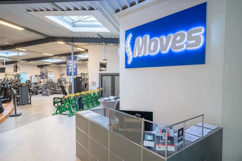 Innenraum eines Fitnessstudios mit Empfangstresen, "Moves"-Schild, Fitnessgeräten und Jubiläumsbanner.